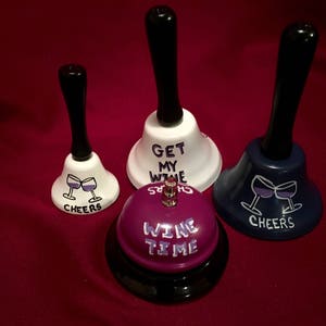May include: Four different hand bells with various wine-themed sayings. The bells are white, purple, and blue with black handles. The sayings on the bells are "Get My Wine", "Cheers", "Cheers", and "Wine Time".