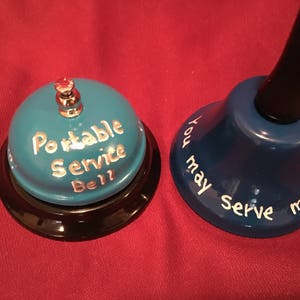 May include: Two decorative bells. One is teal with the words "Portable Service Bell" in white. The other is blue with the words "You may serve me now" in white. Both bells have black bases and are on a red surface.