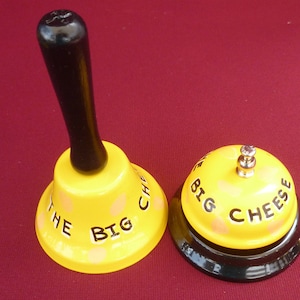 May include: Two yellow hand bells with black lettering. The larger bell has a black handle and reads "The Big Cheese". The smaller bell reads "Big Cheese".