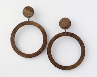 Puede incluir: Un par de pendientes de aro de madera. Cada pendiente presenta un pequeño perno circular conectado a un aro circular grande. Los pendientes están hechos de madera marrón oscuro con vetas visibles.