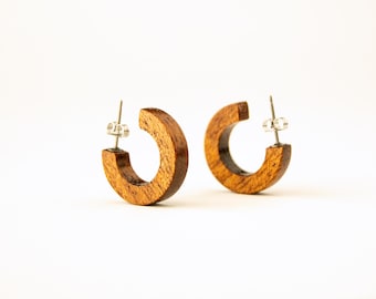 Puede incluir: Un par de pendientes de aro de madera con un acabado marrón natural. Los pendientes están hechos de una pieza gruesa y redondeada de madera y tienen un diseño simple y minimalista.