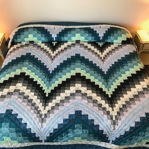 May include: A teal, blue, green, black, and white quilt with a chevron pattern. The quilt is folded in half and is on a bed.