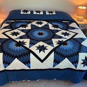 May include: A blue and white quilt with a star pattern. The quilt is made with a variety of shades of blue and white fabric. The quilt is on a bed with a white headboard and a white nightstand.