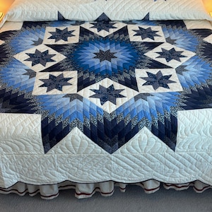May include: A white quilt with a blue and black star pattern. The quilt is on a bed with a white headboard and a white bed skirt.