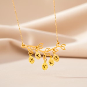 May include: A gold-coloured necklace featuring a branch design with leaf pendants. Each leaf pendant has a single initial letter. The necklace is suspended by a delicate chain. The background is a soft, neutral colour.