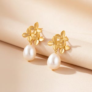 Peut inclure: Boucles d'oreilles dorées avec un motif floral à quatre pétales. Chaque boucle d'oreille comporte une grosse perle blanche en forme de goutte suspendue en dessous. Les boucles d'oreilles sont présentées sur un fond neutre et doux.