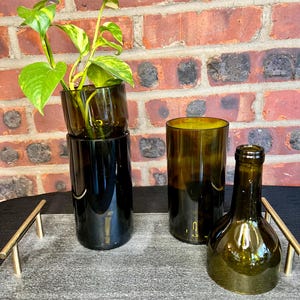 May include: Three glass bottles, two with a dark green tint and one with a clear green tint, are arranged on a gray tray with gold handles. A small green plant is growing in the top of the tallest bottle.