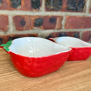May include: Two ceramic strawberry-shaped bowls. The bowls are red with white interiors and green stems. The red exterior has a textured pattern to resemble a strawberry. The bowls are on a wooden surface with a brick background.