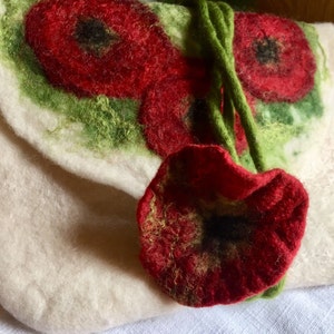 May include: A white felt clutch bag with a green stem and red poppy flower design. The bag has a long green felt strap.