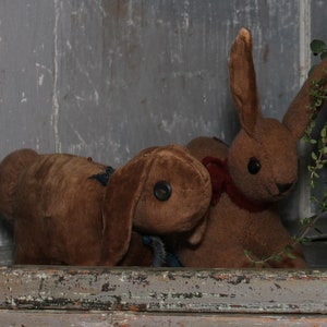May include: Three brown plush bunny rabbits, one with a red ribbon around its neck, sitting on a wooden shelf with a potted plant in the background.