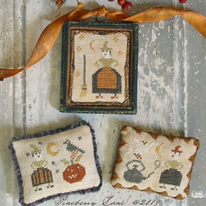 May include: Three cross-stitch embroidery pieces with a Halloween theme. The first piece is framed and features a witch with a broom and stars. The second piece features a witch with a cat and a pumpkin. The third piece features two witches with a cauldron and a moon.