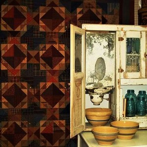 May include: A vintage white cabinet with glass doors and a flour sifter inside. The cabinet has a distressed finish and is decorated with a patchwork quilt.