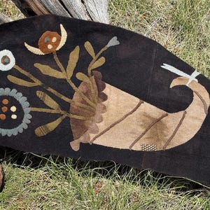 May include: A dark brown, handcrafted textile with a horn of plenty design. The textile features floral and leaf accents in shades of brown, tan, and green. A white and gray accent is on the right side. The textile is placed on a wooden surface.