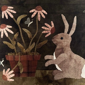 May include: A brown rabbit sits near a potted plant with pink flowers and green leaves. The background is a dark green fabric. There are two butterflies on the fabric.