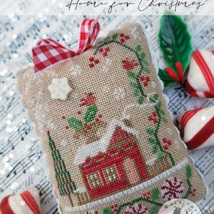 May include: A cross-stitched Christmas ornament with a red house, green trees, and snowflake details. The ornament has a red and white gingham ribbon and the text "SOUVENIRS OF THE HEART Home for Christmas".