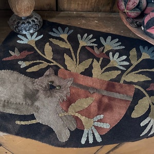 May include: A dark brown, oval-shaped rug with a cat and floral design. The cat is gray with brass-colored eyes, and the flowers are blue, green, and brown. The rug is likely handmade and adds a rustic touch.