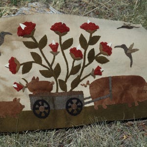 May include: A beige textile art piece featuring a whimsical farm scene. A large pig pulls a cart with two smaller pigs and a floral arrangement of red roses. Hummingbirds and additional roses are also present.