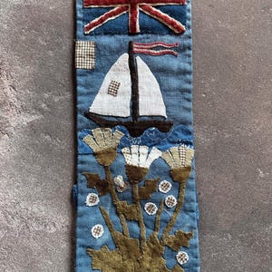 May include: A blue fabric bookmark with a Union Jack flag, sailboat, and floral design. The sailboat has a white sail and is on blue waves. The flowers are brown and white. The bookmark is rectangular and has a button closure.