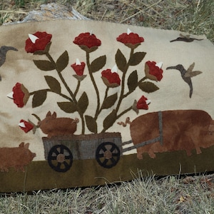 May include: A beige textile art piece featuring a whimsical farm scene. A large pig pulls a cart with two smaller pigs and a floral arrangement of red roses. Hummingbirds and additional roses are also present.