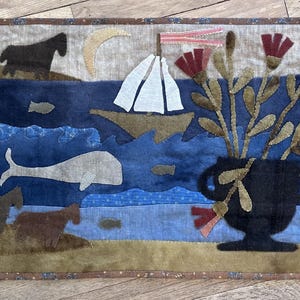 May include: A blue and brown felt wall hanging with a sailboat, whale, fish, and flowers in a vase. The background is a light brown with a crescent moon.