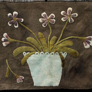 May include: A felted floral design featuring a light blue vase filled with green leaves and purple and white flowers. The artwork is set against a brown background, with a detached flower on the left side.