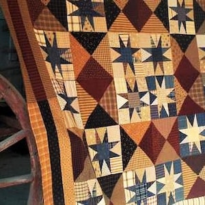 Może przedstawiać: Kołdra w patchworku z różnymi tkaninami w kolorze brązowym, beżowym, niebieskim i białym. Kołdra składa się z kwadratów z motywem gwiazdy w środku każdego kwadratu. Kołdra jest zarzucona na drewniane koło wozu.