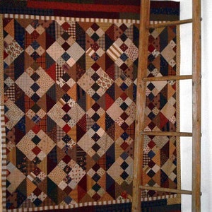 May include: A patchwork quilt featuring a range of colours and patterns, including red, brown, blue, and white. The quilt is hanging on a wall beside a wooden ladder.
