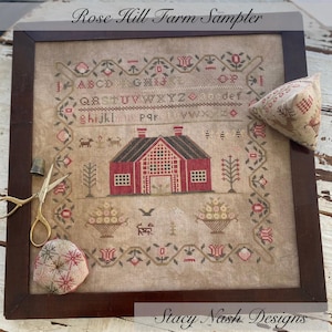 May include: A framed cross-stitch sampler titled "Rose Hill Farm Sampler" by Stacy Nash Designs. The sampler features a red farmhouse with a brown roof, a basket of fruit, and a variety of flowers and plants. The alphabet is stitched in the top portion of the sampler.