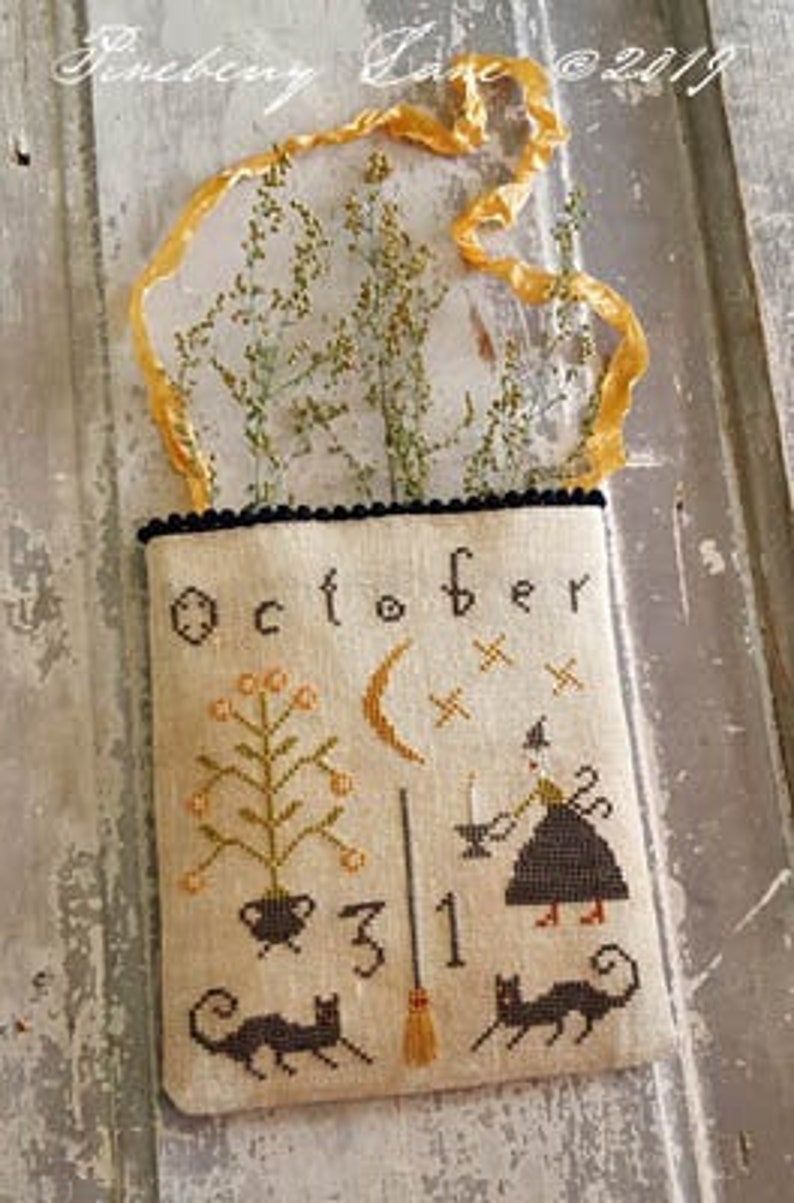 May include: A cross-stitch sampler with the word "October" and a witch with a cauldron, a broom, and a crescent moon. The sampler is framed in a burlap bag with a black trim and a yellow ribbon.