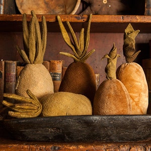 May include: A collection of five felt pineapples in a wooden bowl. The pineapples are in shades of brown and tan, with green felt leaves. The bowl is dark brown and has a rustic look.