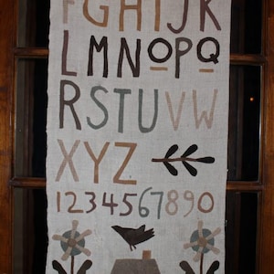 May include: A beige fabric banner with the alphabet, numbers 1-9 and 0, a stylized twig, and two brown silhouettes of flowers with a brown house in the center. The letters and numbers are in brown, green, and beige.