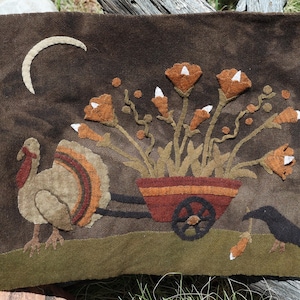 May include: A brown felted wall hanging featuring a turkey, a crow, and a cart filled with flowers. The turkey has a tan body with red, orange, and brown tail feathers. The crow holds a carrot. A crescent moon is in the upper left.