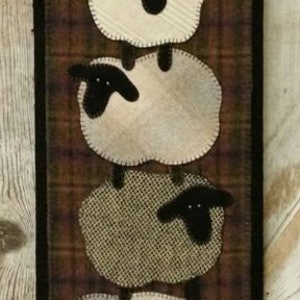 May include: A vertical wall hanging featuring five stacked sheep appliqués on a dark brown plaid background. Each sheep has a black head and a textured body in varying shades of cream, beige, and grey. The bottom edge has five black tabs.