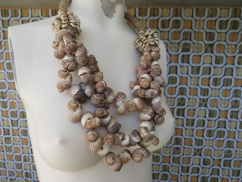 K&ouml;nnte beinhalten: Eine mehrreihige Halskette aus braunen und wei&szlig;en Muscheln. Die Halskette befindet sich auf einer wei&szlig;en Mannequin-B&uuml;ste.