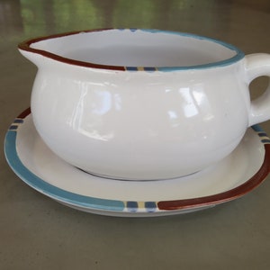May include: White ceramic gravy boat with a blue and brown trim. The boat is sitting on a matching saucer.