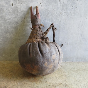 May include: A rustic, handcrafted gourd-shaped vessel with a wooden handle and a woven neck. The top features a carved wooden element with two upward-pointing projections. The vessel is brown and tan in color.