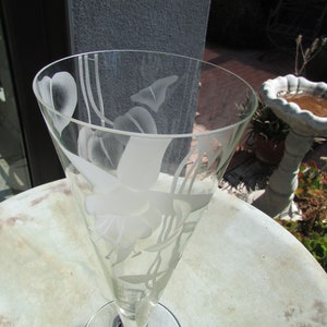 Puede incluir: Una copa de champ&aacute;n de cristal transparente con dise&ntilde;os florales grabados. El vaso tiene una parte superior acampanada y un tallo delgado. El dise&ntilde;o grabado incluye flores y hojas. La copa est&aacute; sobre una mesa redonda de color claro.