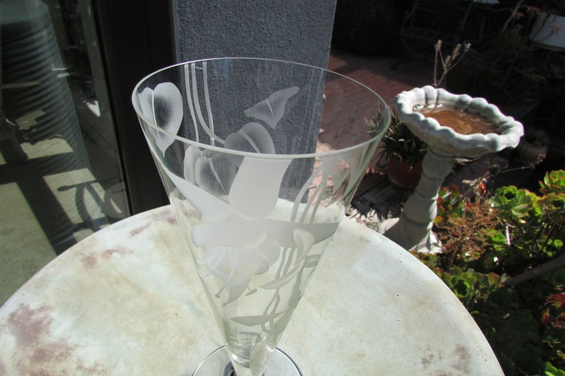 Puede incluir: Una copa de champ&aacute;n de cristal transparente con dise&ntilde;os florales grabados. El vaso tiene una parte superior acampanada y un tallo delgado. El dise&ntilde;o grabado incluye flores y hojas. La copa est&aacute; sobre una mesa redonda de color claro.