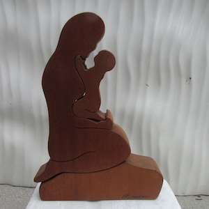 Puede incluir: Una escultura de madera de una madre y un niño, tallada en estilo silueta. La madre sostiene al niño en sus brazos y ambos miran hacia abajo. La escultura está pintada en un color marrón oscuro.
