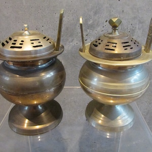 May include: Two antique brass incense burners with intricate designs and lids. The burners have a round base and a cylindrical body with a decorative top.