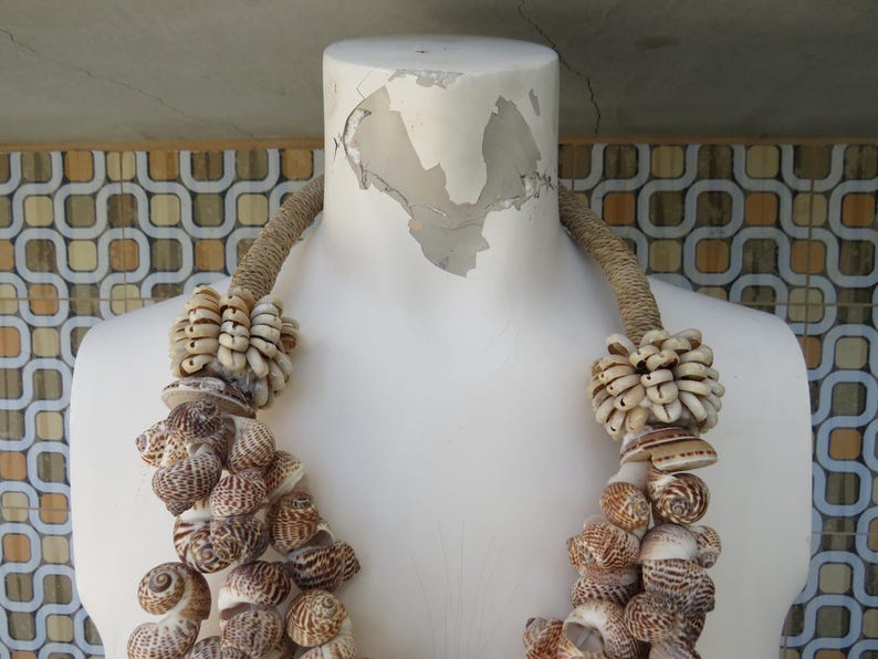 K&ouml;nnte beinhalten: Eine wei&szlig;e Schaufensterpuppe tr&auml;gt eine Halskette aus nat&uuml;rlichen Muscheln. Die Halskette besteht aus zwei Reihen von Muscheln, eine mit gr&ouml;&szlig;eren, braunen Muscheln und die andere mit kleineren, wei&szlig;en Muscheln. Die Halskette ist mit einem braunen Seil gebunden.