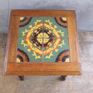 May include: A square wooden side table featuring a tabletop inlaid with four ceramic tiles. The tiles display a symmetrical pattern in shades of green, yellow, orange, and dark brown, framed by a natural wood border with a visible grain texture.