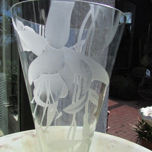 Puede incluir: Un jarr&oacute;n de vidrio transparente con la parte superior acampanada, grabado con un delicado dise&ntilde;o floral. Las flores y hojas blancas esmeriladas crean un hermoso contraste con el vidrio transparente. El jarr&oacute;n probablemente se utiliza con fines decorativos.
