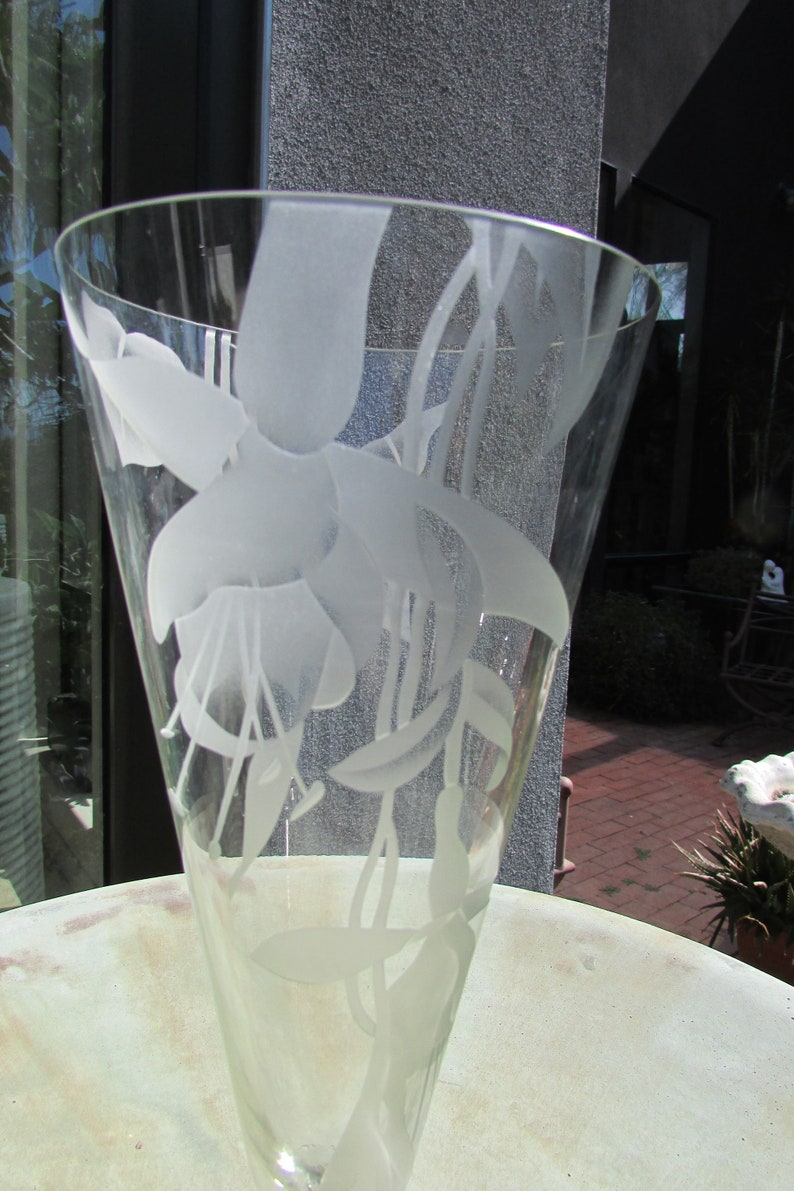 Puede incluir: Un jarr&oacute;n de vidrio transparente con la parte superior acampanada, grabado con un delicado dise&ntilde;o floral. Las flores y hojas blancas esmeriladas crean un hermoso contraste con el vidrio transparente. El jarr&oacute;n probablemente se utiliza con fines decorativos.