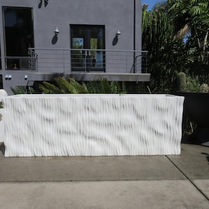 May include: A large, white, rectangular planter with a wavy, textured surface. The planter is made of a smooth, white material and has a modern design.