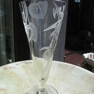 Puede incluir: Un jarr&oacute;n de cristal transparente con la parte superior acampanada y una base redonda. El jarr&oacute;n est&aacute; grabado con dise&ntilde;os florales, incluyendo hojas y flores. El jarr&oacute;n est&aacute; sobre una mesa redonda de color claro.