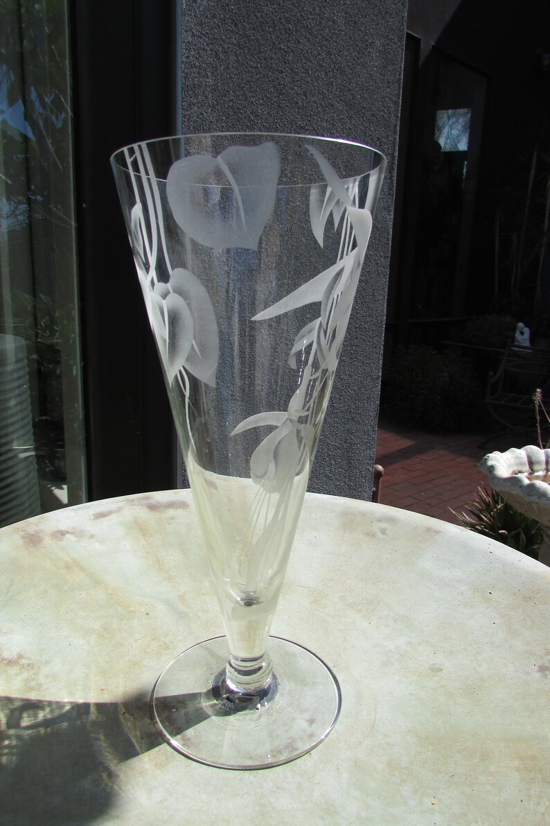 Puede incluir: Un jarr&oacute;n de cristal transparente con la parte superior acampanada y una base redonda. El jarr&oacute;n est&aacute; grabado con dise&ntilde;os florales, incluyendo hojas y flores. El jarr&oacute;n est&aacute; sobre una mesa redonda de color claro.