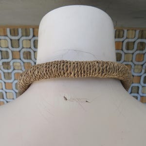 K&ouml;nnte beinhalten: Nahaufnahme einer braunen geflochtenen Halskette auf einer wei&szlig;en Puppe. Die Halskette ist aus Naturfasern gefertigt und hat ein schlichtes, rustikales Design.