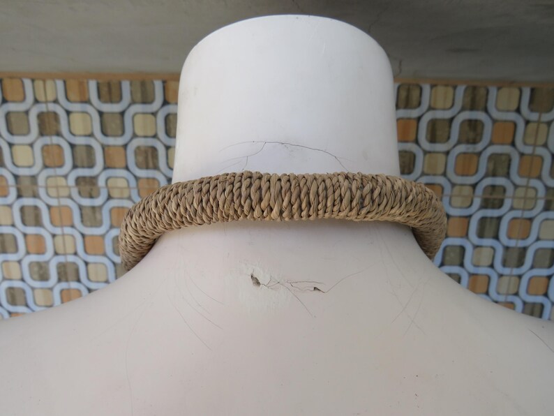 K&ouml;nnte beinhalten: Nahaufnahme einer braunen geflochtenen Halskette auf einer wei&szlig;en Puppe. Die Halskette ist aus Naturfasern gefertigt und hat ein schlichtes, rustikales Design.