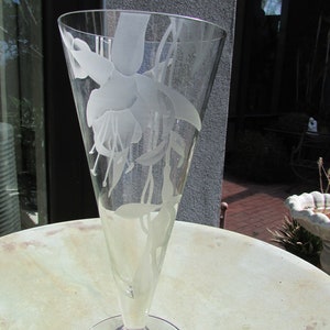 Puede incluir: Un jarr&oacute;n de cristal transparente con la parte superior acampanada y una base redonda. El jarr&oacute;n est&aacute; grabado con un dise&ntilde;o floral con flores y hojas de fucsia. El jarr&oacute;n est&aacute; sobre una mesa redonda.